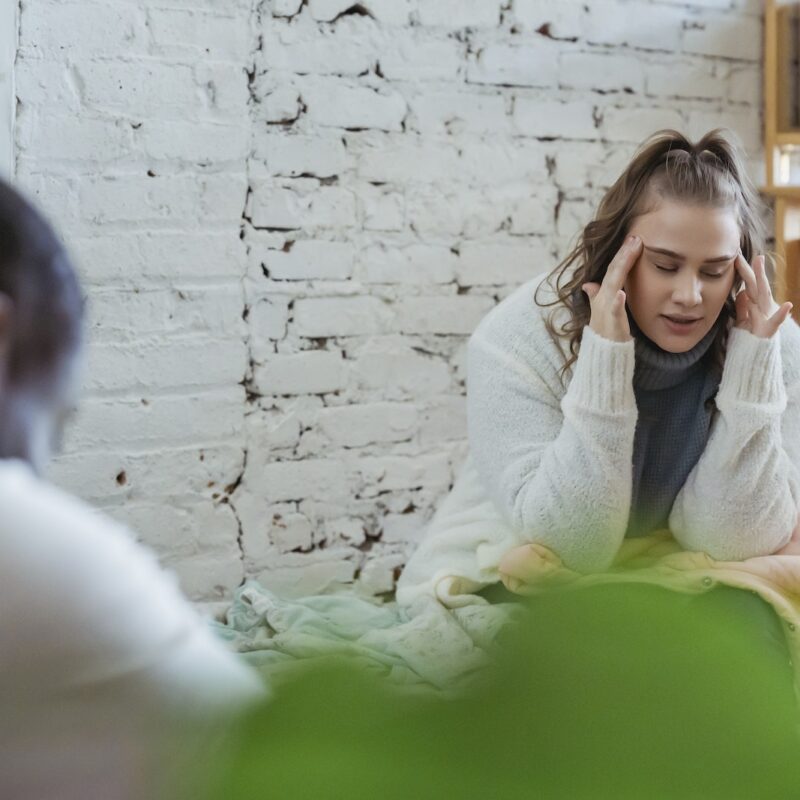 Exhausted woman calming down after argument with husband by putting fingers on temples and man sitting and looking down